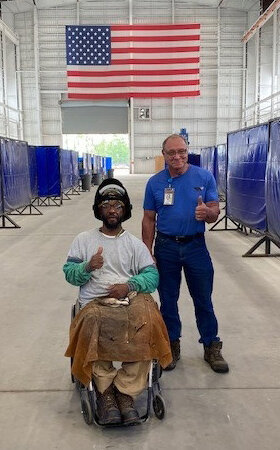 Larry Phillips, a Black man who uses a wheelchair, posing for a photo in a work setting against the backdrop of the American flag with another man, as both give thumbs-up symbols.